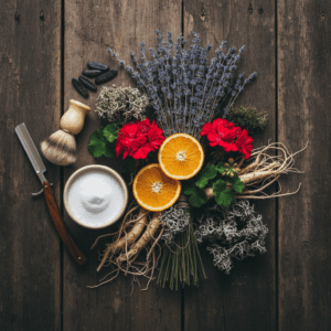 Flat lay photography on a rustic wood surface, illustrating the key ingredients of the Fougère perfume accord: dried lavender, bergamot, geranium, vetiver roots, oakmoss, and tonka beans. They are arranged next to a vintage shaving brush, a foaming shaving bowl, and a straight razor, evoking the traditional barbershop universe.