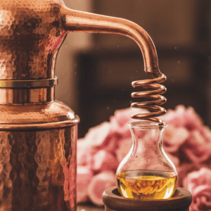 Detailed photography of a traditional copper alembic and a Florentine flask separating essential oil from floral water, with rose petals in the background.