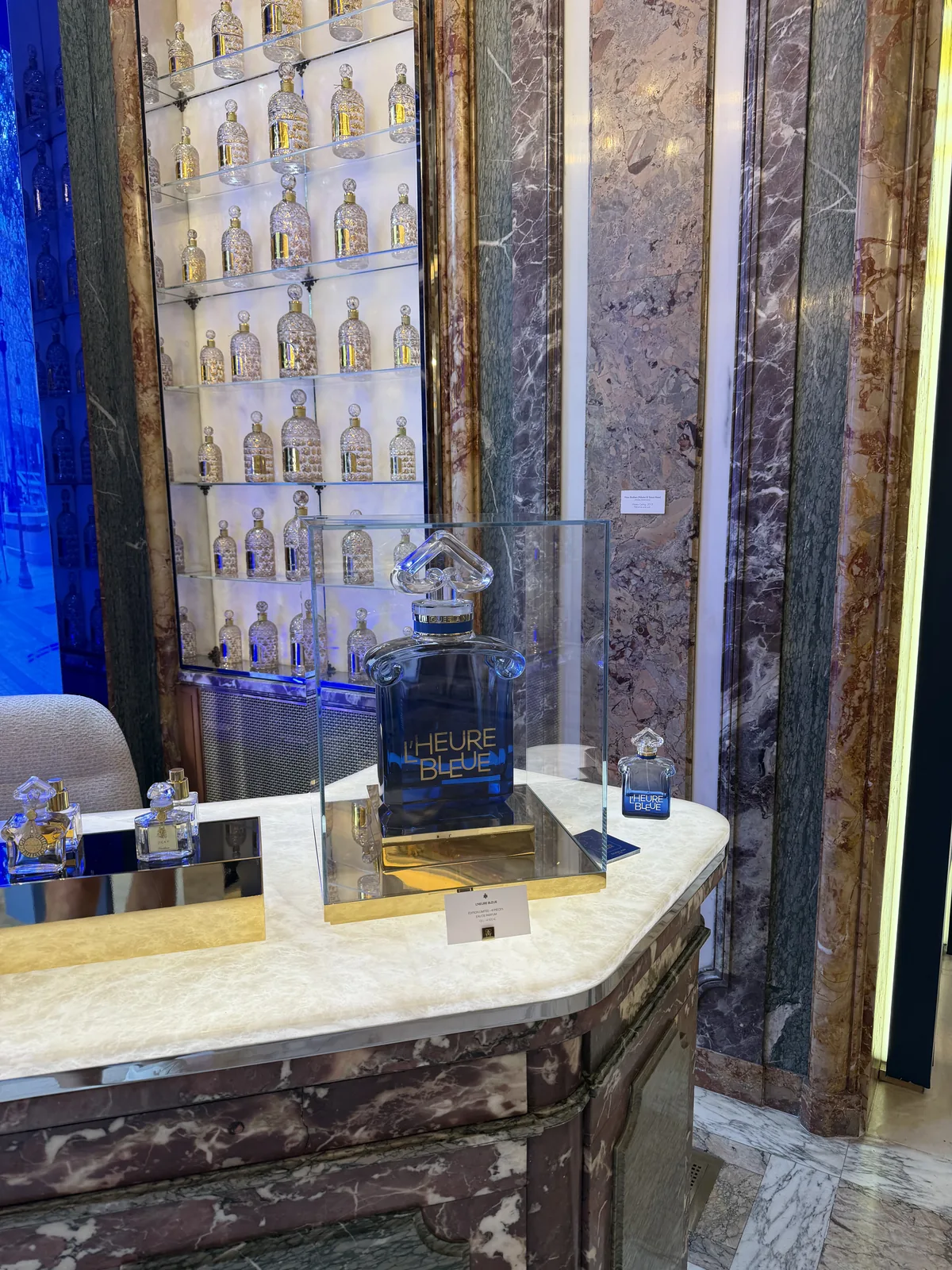 Flacon géant de L'Heure Bleue sous vitrine devant le mur de flacons abeilles Guerlain.
