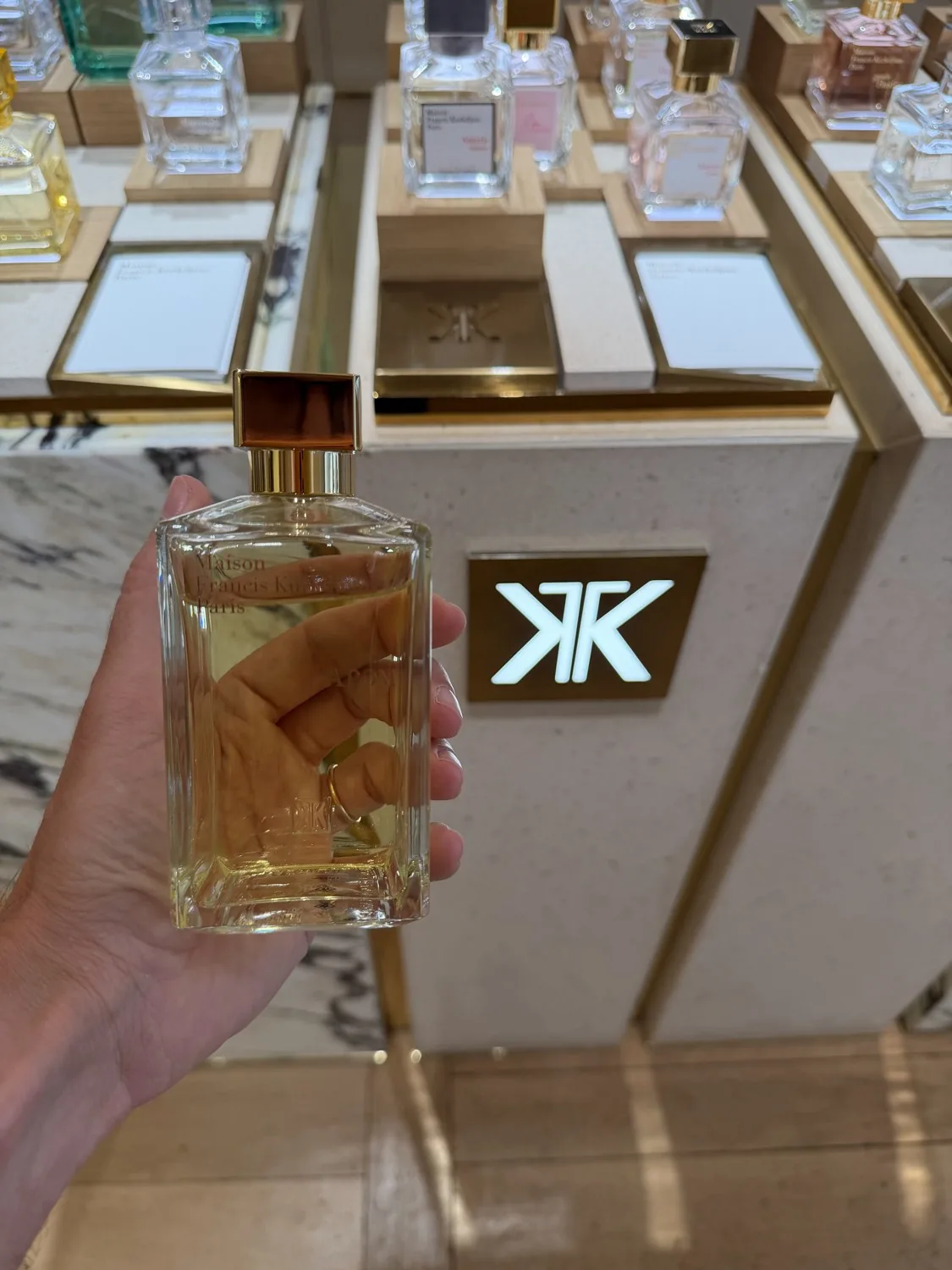 Flacon d'APOM de Maison Francis Kurkdjian tenu en main devant le logo de la Maison.