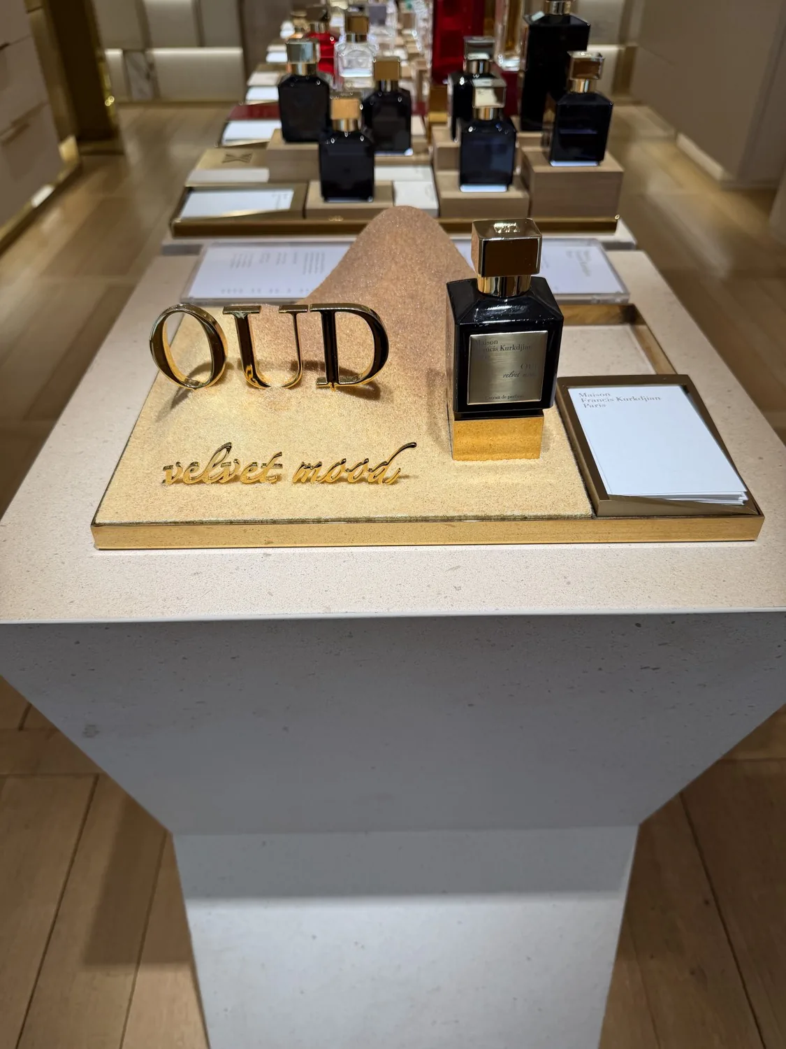 Présentoir Oud Velvet Mood de Maison Francis Kurkdjian, flacon noir et or posé sur du sable.