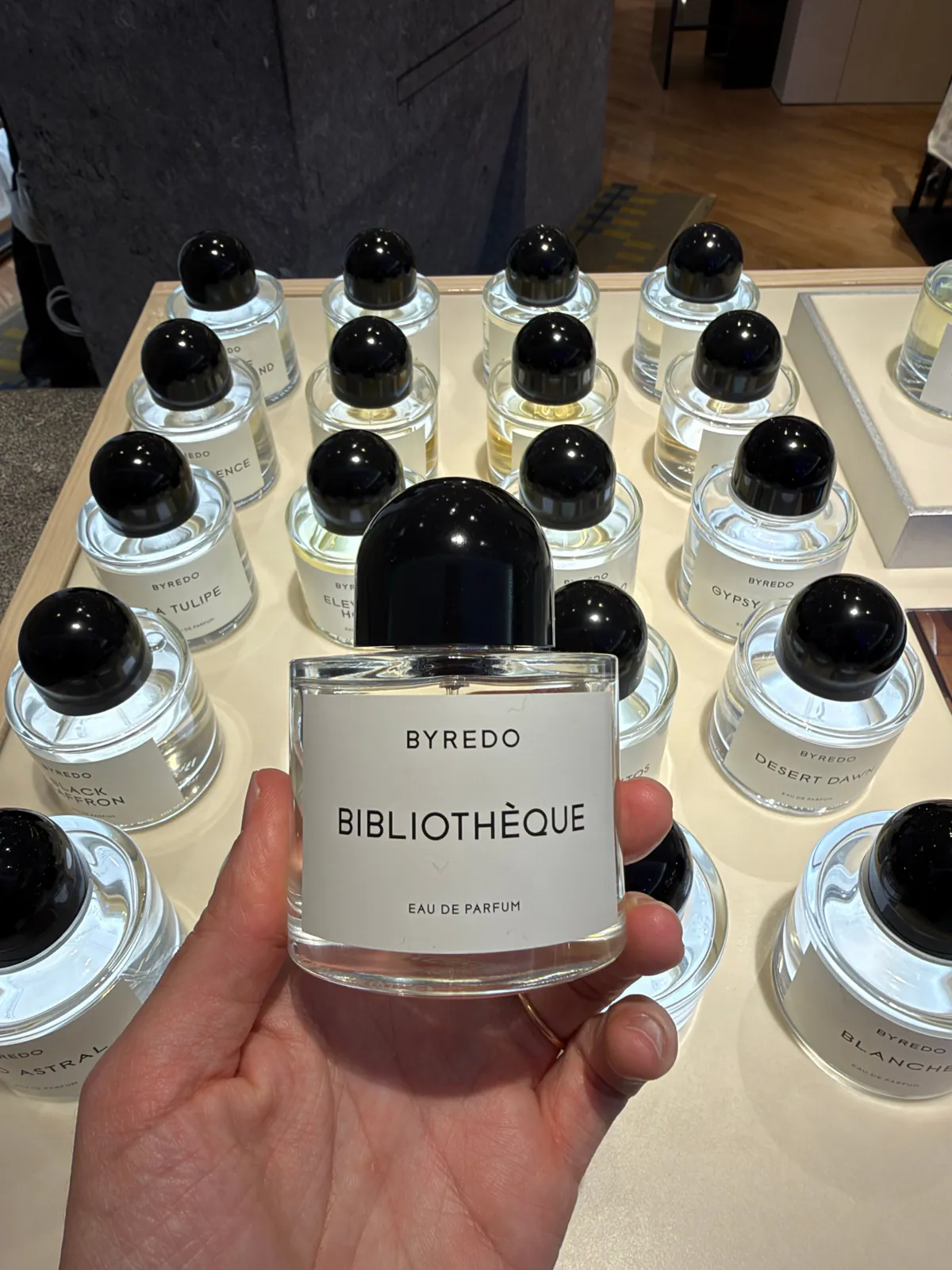 Flacon d'eau de parfum Bibliothèque de Byredo tenu en main devant les autres parfums de la marque.