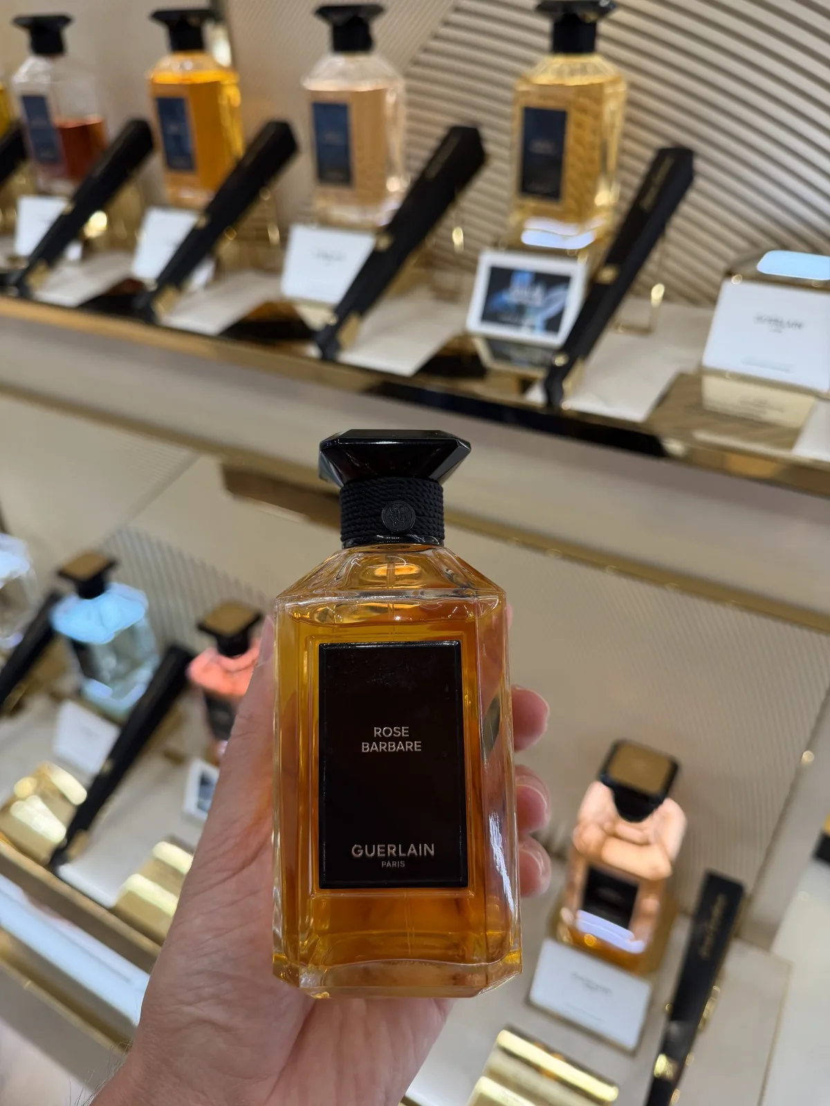 Flakon von Rose Barbare von Guerlain in der Hand, vor der Wand der Kollektion L'Art et la Matière.