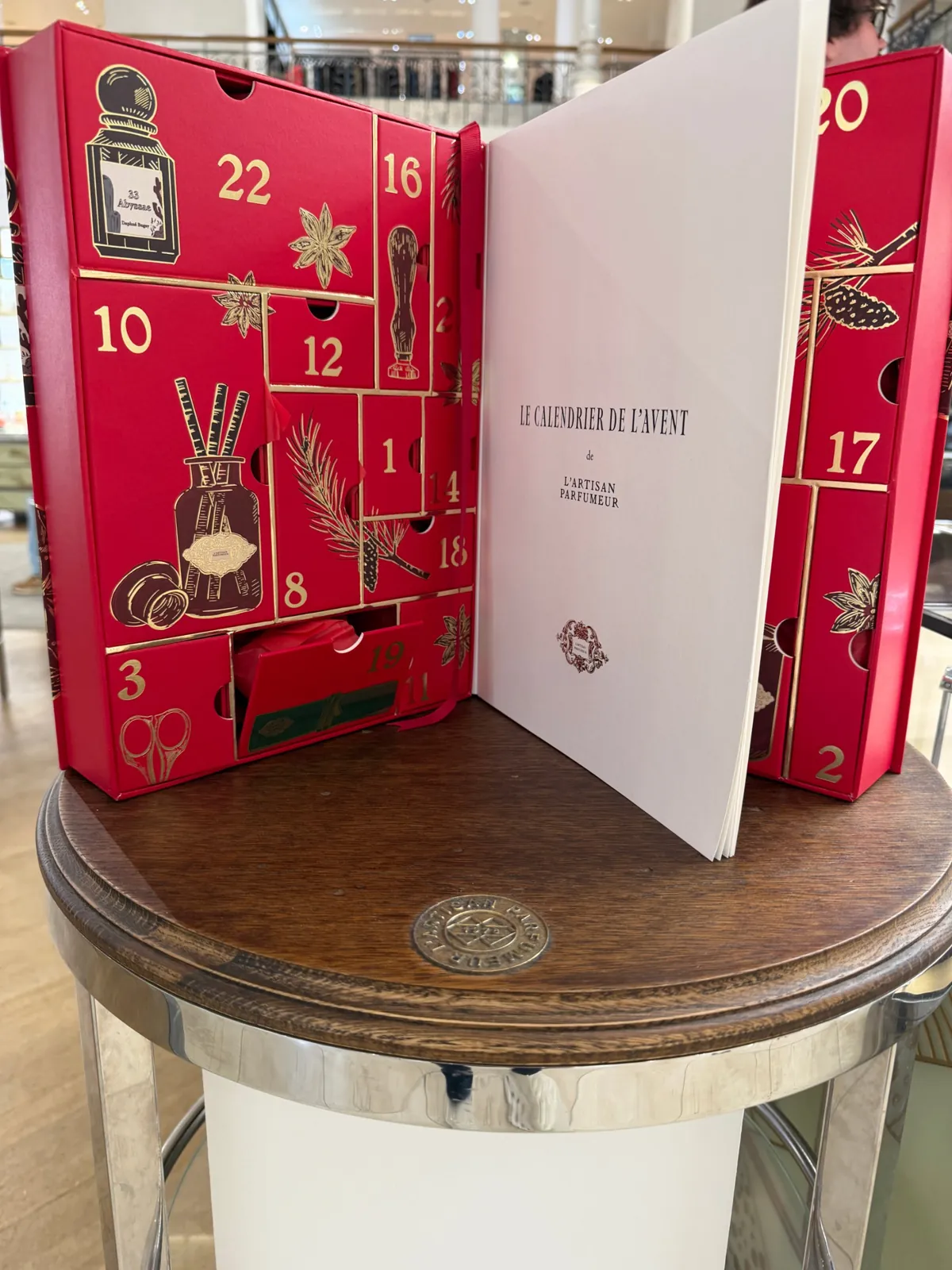 Calendrier de l'Avent rouge et or de L'Artisan Parfumeur, ouvert sur son livret.